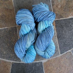 May include: Two skeins of blue yarn in varying shades of light and dark blue. The yarn is thick and appears hand-dyed, with a soft, textured appearance. The yarn is arranged on a stone tile surface.