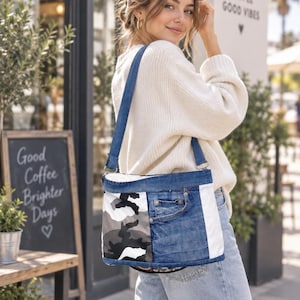 Pode incluir: Uma bolsa de ombro feita de tecido jeans azul, apresentando um painel com padrão de camuflagem cinza e branco no lado esquerdo, uma seção central com detalhe de bolso jeans e uma faixa vertical branca no lado direito com alça azul.