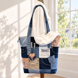 May include: A patchwork tote bag constructed from various shades of blue denim and tan fabric, featuring a white rectangular label with black text, a zipper closure, and a small heart-shaped charm hanging from a metal ring on the front strap.