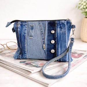 Puede incluir: Un bolso de mano de mezclilla hecho de jeans reciclados. La bolsa presenta un cierre de cremallera, tres botones plateados y una correa de muñeca. El diseño de patchwork incorpora varios tonos de mezclilla azul, creando un accesorio único y elegante.