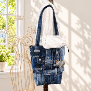 May include: A tote bag constructed from various shades of blue denim fabric patches, featuring visible seams, frayed edges, and metal rivets. A small, heart-shaped fabric charm with a white floral pattern hangs from the side of the bag.