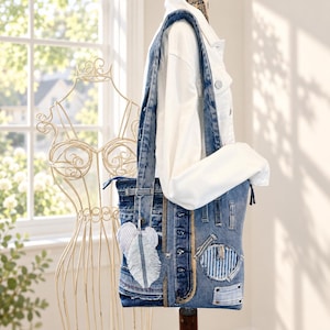 Puede incluir: Un bolso de hombro confeccionado con tela de mezclilla azul reutilizada, con una tapeta de botones vertical, múltiples bolsillos y bordes deshilachados. Un dije de pluma de tela a rayas blancas y azules cuelga de la correa, también de mezclilla.