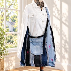 May include: A large hobo-style shoulder bag constructed from repurposed denim fabric in varying shades of blue and black. The bag features a prominent external pocket, frayed edges, and small, scattered pink embroidered accents throughout.