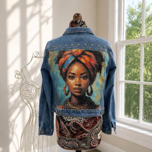 May include: A blue denim jacket featuring a large, colorful graphic print on the back. The print depicts a person wearing a vibrant headwrap, hoop earrings, and a beaded necklace. The jacket includes a collar, long sleeves, and metal studs.