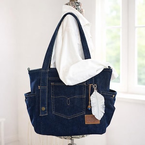 May include: A dark blue denim tote bag with multiple pockets and a light-coloured feather charm. The bag has two handles and a pocket on the front. A white shirt is draped over the bag.