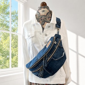 Op de afbeelding: Een blauwe crossbodytas van denim met meerdere ritsvakken, bronskleurige hardware en een rechthoekig stoffen label. De tas wordt gedragen over een wit spijkerjack met een sjaal met patroon zichtbaar bij de halslijn.