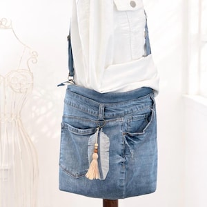 Könnte beinhalten: Hellblaue Jeans-Umhängetasche aus recycelten Jeans. Die Tasche hat die originalen Taschen und Gürtelschlaufen. Ein dekorativer Federanhänger mit Holzperlen und Quaste hängt am Reißverschluss.