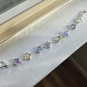 May include: A silver bracelet featuring star-shaped charms, each containing a different colored floral element. The bracelet has a lobster clasp closure. The colors include purple, yellow, and blue. The bracelet is displayed on a white surface.