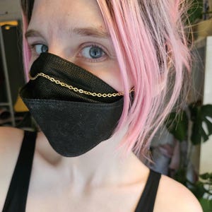 May include: A person wearing a black face mask with a gold chain attached to the mask. The person has pink hair.