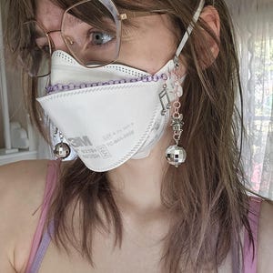 May include: A white N95 mask with a purple chain and decorative charms, including a musical note and a disco ball. The mask is worn with glasses and a pink top. The mask has the text "3M" and "NIOSH".