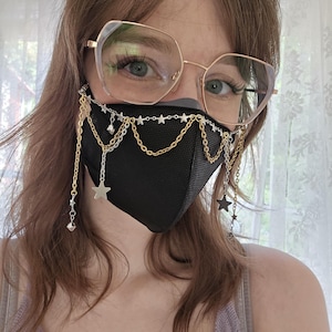 Puede incluir: Una mascarilla negra adornada con cadenas doradas y plateadas, estrellas y cristales colgantes. La mascarilla se lleva con gafas con montura dorada. Las cadenas están drapeadas sobre la mascarilla y cuelgan, creando un efecto decorativo. La estética general es una mezcla de practicidad y moda.