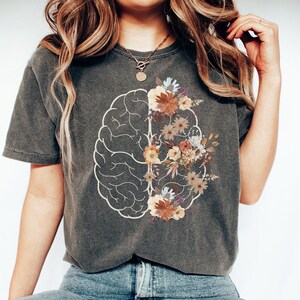 May include: A dark gray t-shirt with a white outline of a brain with floral designs inside. The flowers are in shades of brown, orange, and white.