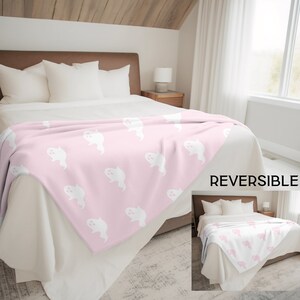 May include: A pink fleece throw blanket with a white ghost pattern. The blanket is reversible, with a solid pink side and a side with the ghost pattern. The blanket is shown on a bed with white bedding.
