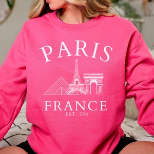 May include: A pink sweatshirt with a white graphic design of the Eiffel Tower, the Arc de Triomphe, and the Louvre Pyramid. The text "PARIS FRANCE EST. 259" is printed below the graphic.