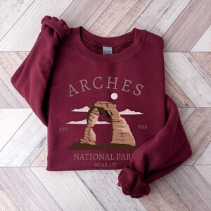 May include: Burgundy sweatshirt with a graphic of Delicate Arch in Arches National Park, Moab, Utah. The text "ARCHES" is above the arch and "EST. 1929" is below the arch.