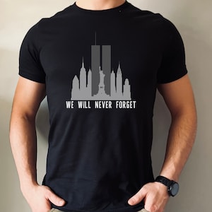May include: Black t-shirt with a gray silhouette of the New York City skyline, including the Twin Towers, the Statue of Liberty, and other buildings. The text "WE WILL NEVER FORGET" is printed below the skyline.
