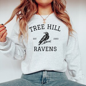 May include: A gray sweatshirt with the text "Tree Hill" and "Ravens" printed on it. The text is in a black font and there is a drawing of a raven in between the words. The text "Est. 2003" is printed below the raven.