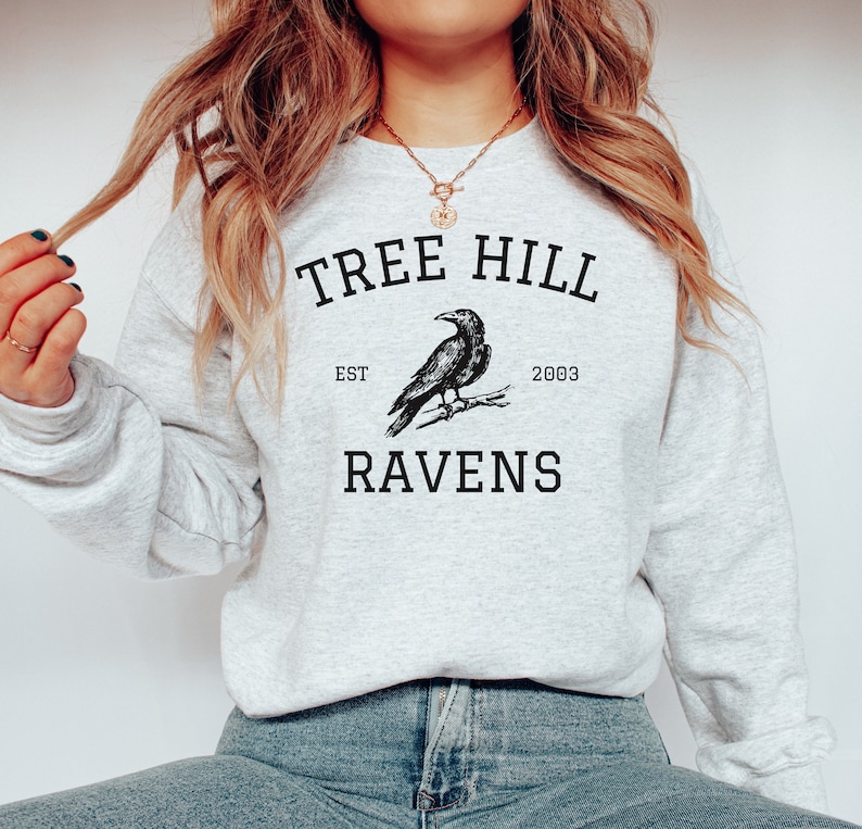 May include: A gray sweatshirt with the text "Tree Hill" and "Ravens" printed on it. The text is in a black font and there is a drawing of a raven in between the words. The text "Est. 2003" is printed below the raven.
