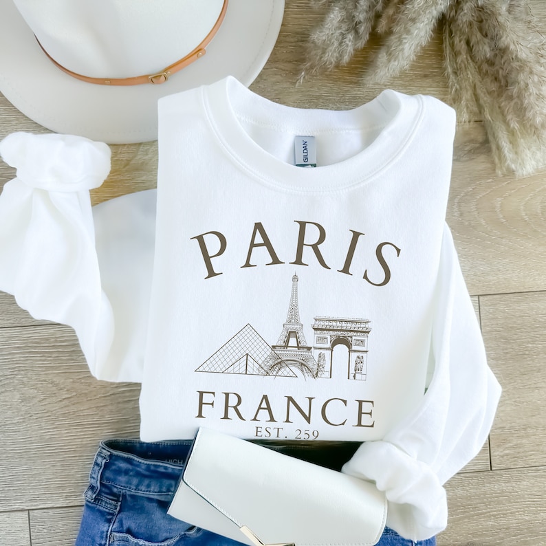 May include: White sweatshirt with a brown graphic print of the Eiffel Tower, the Louvre, and the Arc de Triomphe. The text "PARIS FRANCE EST. 259" is printed below the graphic.