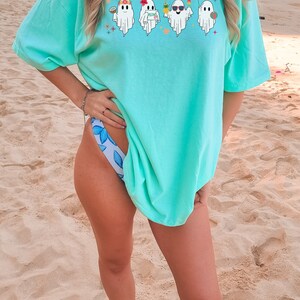 K&ouml;nnte beinhalten: Ein t&uuml;rkisfarbenes Oversize-T-Shirt mit einem Aufdruck von vier Geistern, die Sonnenbrillen, Blumenketten und Getr&auml;nke tragen. Die Geister tragen Blumenkr&auml;nze und haben eine Strand-Atmosph&auml;re. Das T-Shirt wird von einer Person getragen, die auf einem Sandstrand steht.
