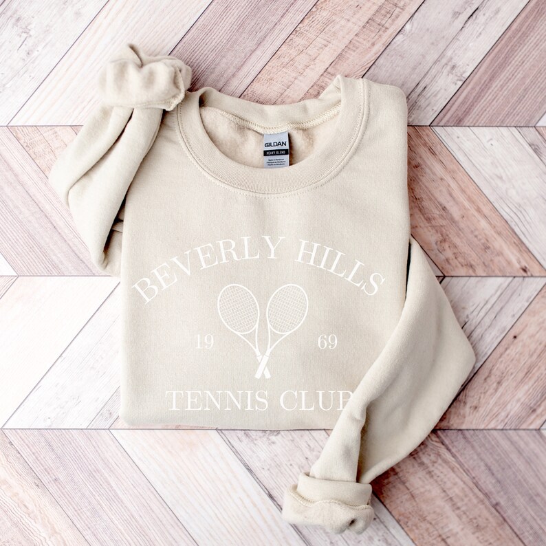 May include: Beige sweatshirt with a white graphic of two tennis rackets crossed over each other with the text "Beverly Hills Tennis Club" and the numbers "19" and "69" on either side of the rackets.