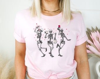May include: Pink t-shirt with a graphic of three skeletons dancing and holding Valentine's Day themed items. The skeletons are wearing black and white and are surrounded by pink hearts.