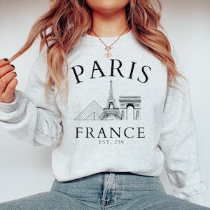 May include: Grey sweatshirt with a black graphic of the Eiffel Tower, the Louvre Pyramid, and the Arc de Triomphe. The text "PARIS FRANCE EST. 259" is printed below the graphic.
