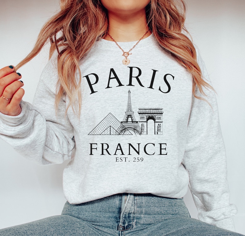 May include: Grey sweatshirt with a black graphic of the Eiffel Tower, the Louvre Pyramid, and the Arc de Triomphe. The text "PARIS FRANCE EST. 259" is printed below the graphic.
