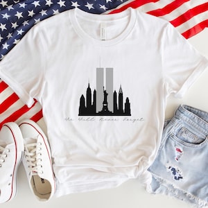May include: White t-shirt with a black silhouette of the New York City skyline, including the Twin Towers, the Statue of Liberty, and other buildings. The text "We Will Never Forget" is written in black cursive below the skyline.