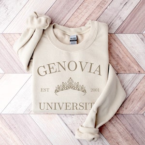 May include: A cream-coloured sweatshirt featuring the text "GENOVIA UNIVERSITY" and "EST 2001" in a darker cream shade. A crown graphic is positioned between the words. The sweatshirt is folded, with the sleeves displayed.