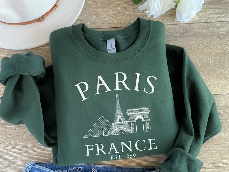 May include: A dark green sweatshirt with a white graphic print of the Eiffel Tower, the Arc de Triomphe, and the Louvre Pyramid. The text "PARIS FRANCE EST. 259" is printed below the graphic.