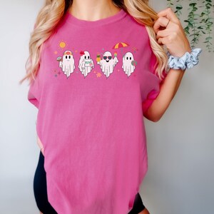 K&ouml;nnte beinhalten: Ein pinkfarbenes T-Shirt mit einem Motiv von vier Geistern, die Sonnenbrillen tragen, Getr&auml;nke und Strandaccessoires halten. Die Geister tragen Blumenkr&auml;nze und haben fr&ouml;hliche Gesichter. Einer der Geister tr&auml;gt den Text "Aloha".