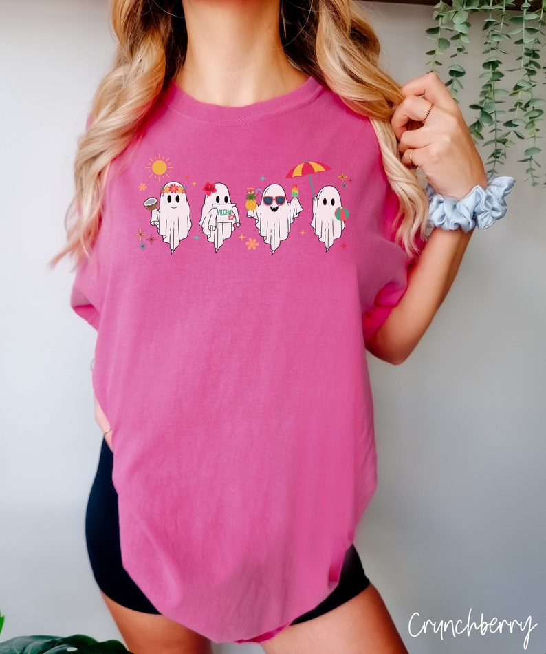 K&ouml;nnte beinhalten: Ein pinkfarbenes T-Shirt mit einem Motiv von vier Geistern, die Sonnenbrillen tragen, Getr&auml;nke und Strandaccessoires halten. Die Geister tragen Blumenkr&auml;nze und haben fr&ouml;hliche Gesichter. Einer der Geister tr&auml;gt den Text "Aloha".