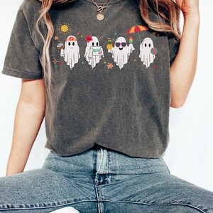 K&ouml;nnte beinhalten: Ein dunkelgraues T-Shirt mit einem Aufdruck von f&uuml;nf Geistern, die Sonnenbrillen tragen, Getr&auml;nke halten und den Strand genie&szlig;en. Die Geister tragen Blumenkr&auml;nze und halten Strandaccessoires wie einen Strandball, eine Ananas und einen Cocktail.