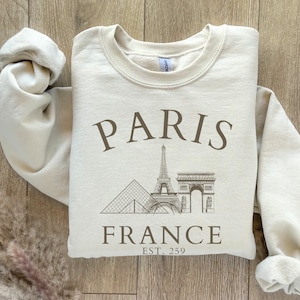 May include: A beige sweatshirt with a graphic print of the Eiffel Tower, the Arc de Triomphe, and the Louvre Pyramid. The text "PARIS FRANCE EST. 259" is printed below the graphic.