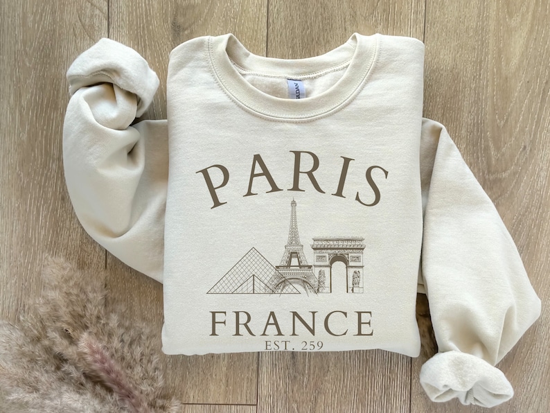 May include: A beige sweatshirt with a graphic print of the Eiffel Tower, the Arc de Triomphe, and the Louvre Pyramid. The text "PARIS FRANCE EST. 259" is printed below the graphic.