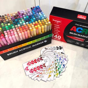 Puede incluir: Un juego de 240 rotuladores de pincel acr&iacute;lico en una caja negra con degradado arco&iacute;ris. La caja est&aacute; etiquetada como "BRUSH ACRYLIC MARKER". Tambi&eacute;n hay una caja roja y negra con "Guang Na" y "ACR". Un abanico de muestras de color est&aacute; en primer plano.