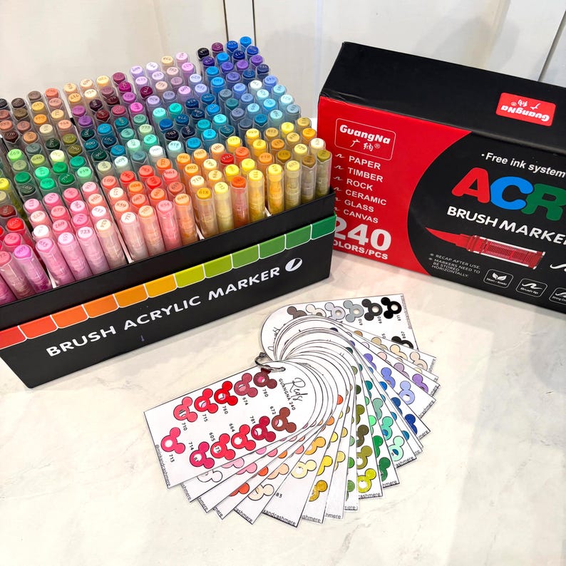 Puede incluir: Un juego de 240 rotuladores de pincel acr&iacute;lico en una caja negra con degradado arco&iacute;ris. La caja est&aacute; etiquetada como "BRUSH ACRYLIC MARKER". Tambi&eacute;n hay una caja roja y negra con "Guang Na" y "ACR". Un abanico de muestras de color est&aacute; en primer plano.