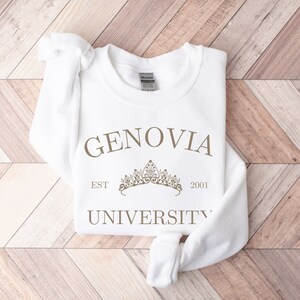 May include: A white sweatshirt featuring the text "GENOVIA UNIVERSITY EST 2001" in a brown font, and a crown graphic. The sweatshirt is folded on a light brown wooden surface.