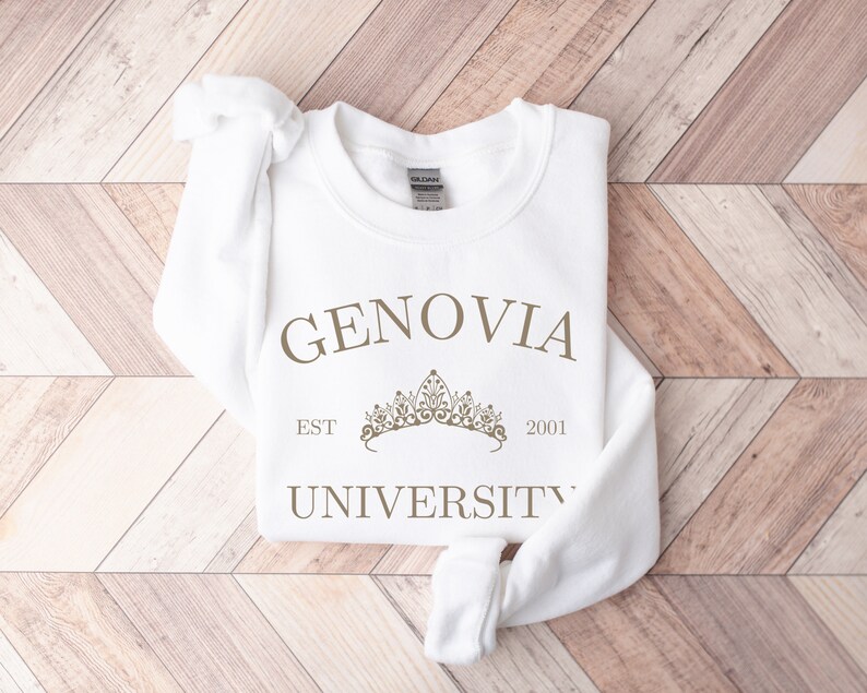 May include: A white sweatshirt featuring the text "GENOVIA UNIVERSITY EST 2001" in a brown font, and a crown graphic. The sweatshirt is folded on a light brown wooden surface.