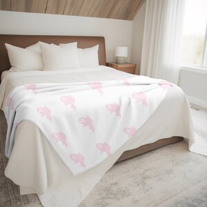 May include: A white fleece throw blanket with a pink ghost pattern. The blanket is folded and placed on a bed with white bedding.