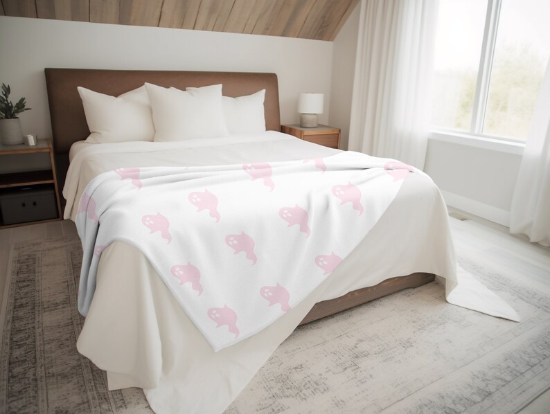 May include: A white fleece throw blanket with a pink ghost pattern. The blanket is folded and placed on a bed with white bedding.
