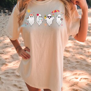 K&ouml;nnte beinhalten: Ein wei&szlig;es T-Shirt mit einem Aufdruck von vier Geistern, die Sonnenbrillen, Blumenketten und Strandutensilien tragen. Die Geister stehen in einer Reihe und haben unterschiedliche Gesichtsausdr&uuml;cke. Einer der Geister h&auml;lt ein Schild mit der Aufschrift "Aloha Day" in der Hand.