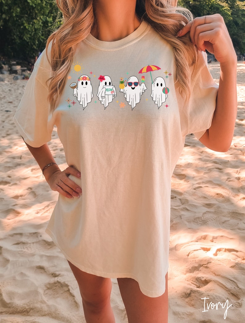 K&ouml;nnte beinhalten: Ein wei&szlig;es T-Shirt mit einem Aufdruck von vier Geistern, die Sonnenbrillen, Blumenketten und Strandutensilien tragen. Die Geister stehen in einer Reihe und haben unterschiedliche Gesichtsausdr&uuml;cke. Einer der Geister h&auml;lt ein Schild mit der Aufschrift "Aloha Day" in der Hand.