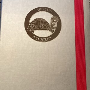 May include: A silver journal with a red elastic band closure. The front cover features a gold embossed design of a turtle with the text "ARE YOU A TURTLE?"