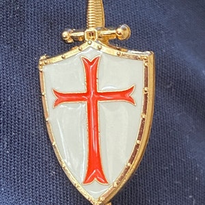 May include: A gold-colored shield with a red cross on a white background. The shield is topped with a sword.