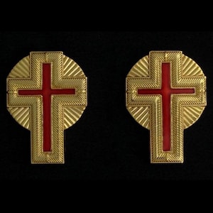 May include: Two gold-colored cross-shaped pins with red centers. The crosses are set against a gold circular background with a sunburst design. The pins are displayed on a black surface.