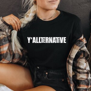 以下が含まれることがあります： 前面に白い文字で「Y'ALLTERNATIVE」とプリントされた黒いTシャツ。長いブロンドの髪とチェックシャツを着た人が着ています。