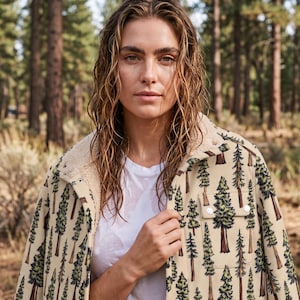 May include: A beige jacket featuring a repeating pattern of green and brown pine trees, a white sherpa fleece lining, a fold-over collar, and white snap buttons. The jacket is worn over a plain white t-shirt with a visible chest pocket.
