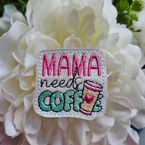 May include: White fabric patch with pink, green and brown embroidery. The text reads "MAMA needs COFFE" with a pink heart on a brown coffee cup.